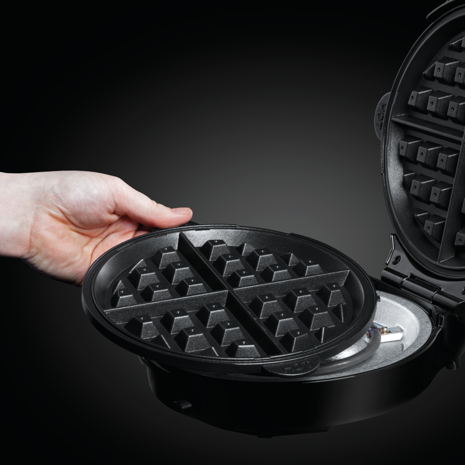 A hand is holding a waffle iron. The iron is open, showing the waffle grids. It's black and on a black background.