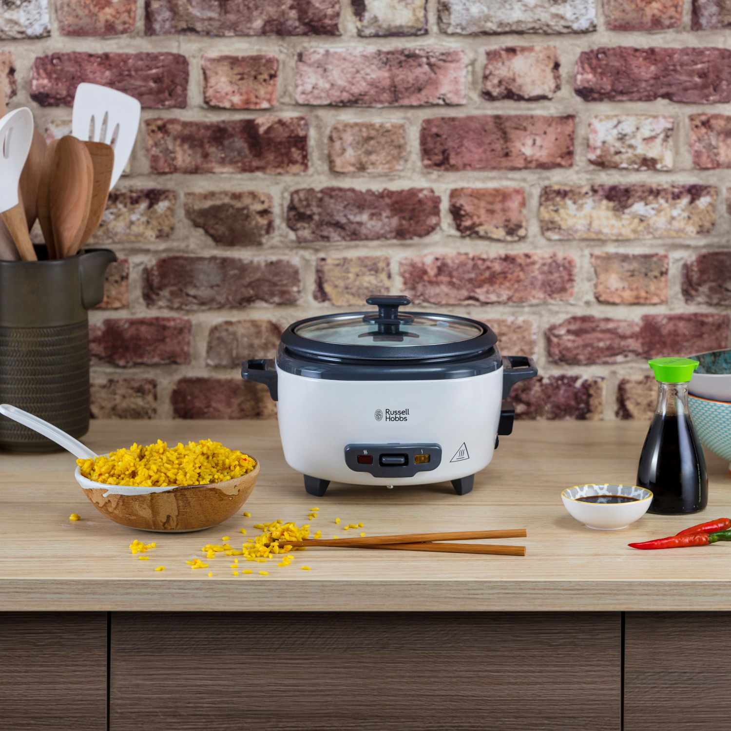 A white rice cooker on a wooden surface, with rice, chopsticks, and soy sauce, against a brick wall.