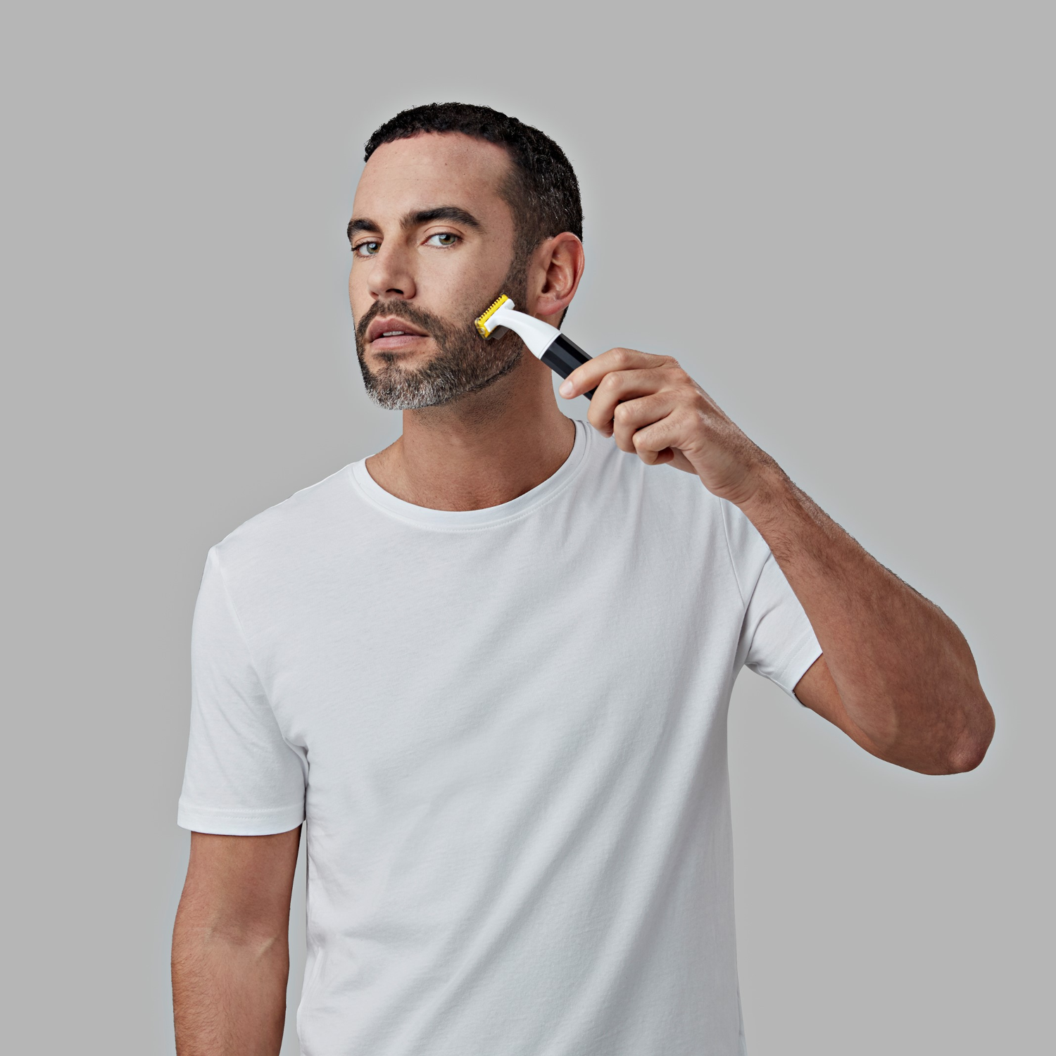A man shaving his beard with a black and white trimmer, against a gray background.