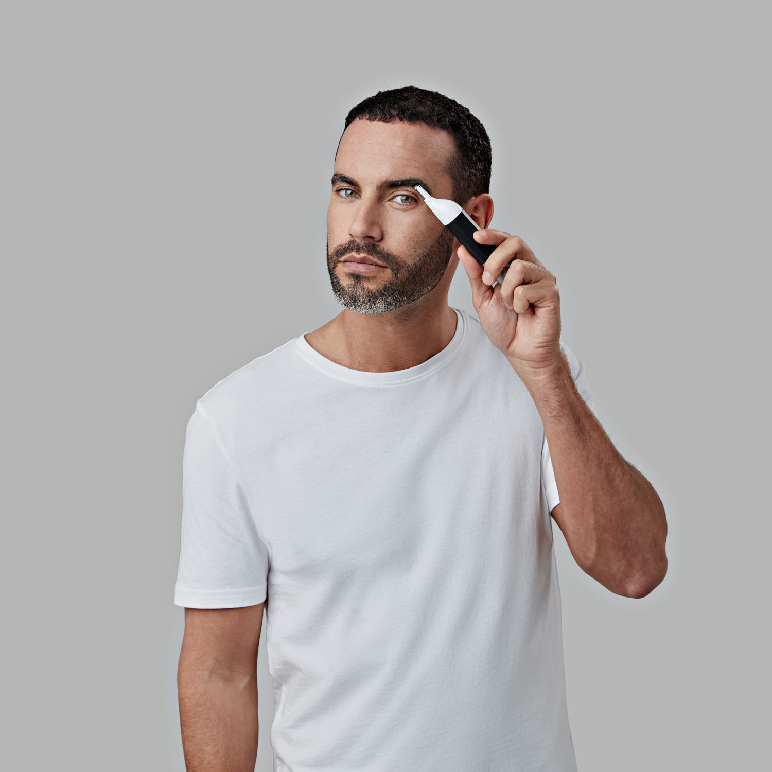 Man using a trimmer on his eyebrow. He has a white tee and gray background.