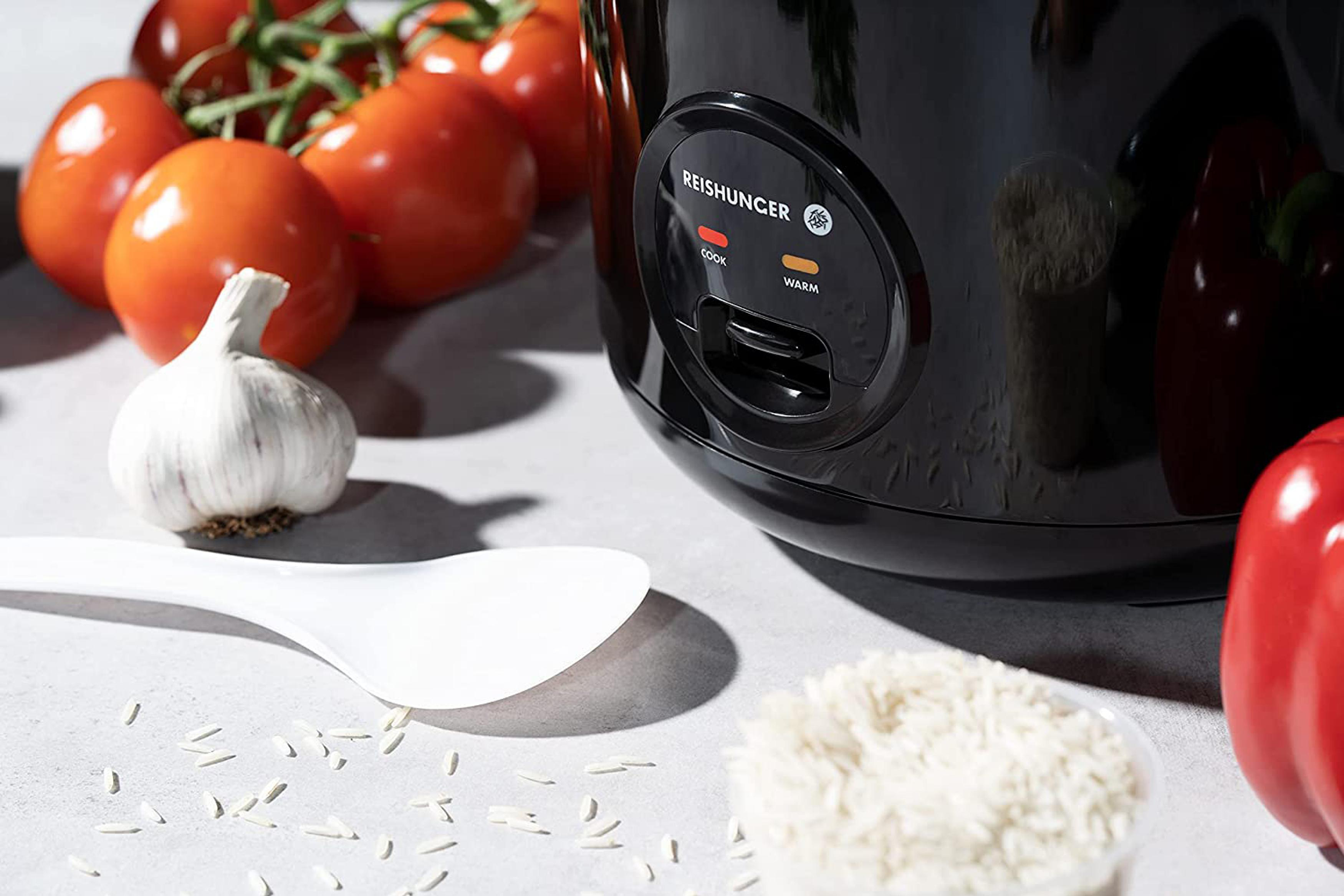 Nahaufnahme eines Reiskochers. Reis, Tomaten, Knoblauch und ein Löffel sind vorhanden.