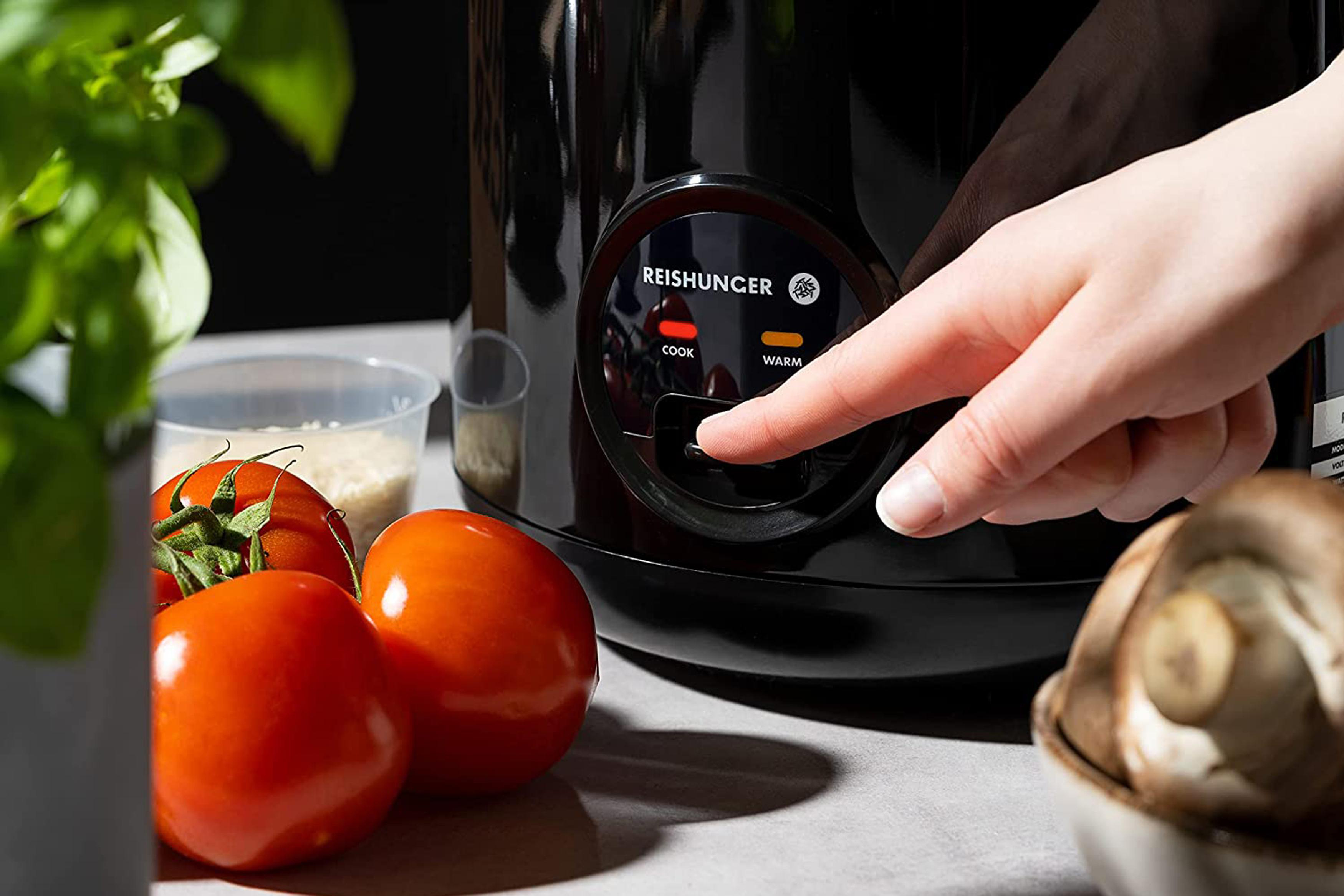 Der Finger einer Person drückt die 'Kochen'-Taste an einem schwarzen Reiskocher.