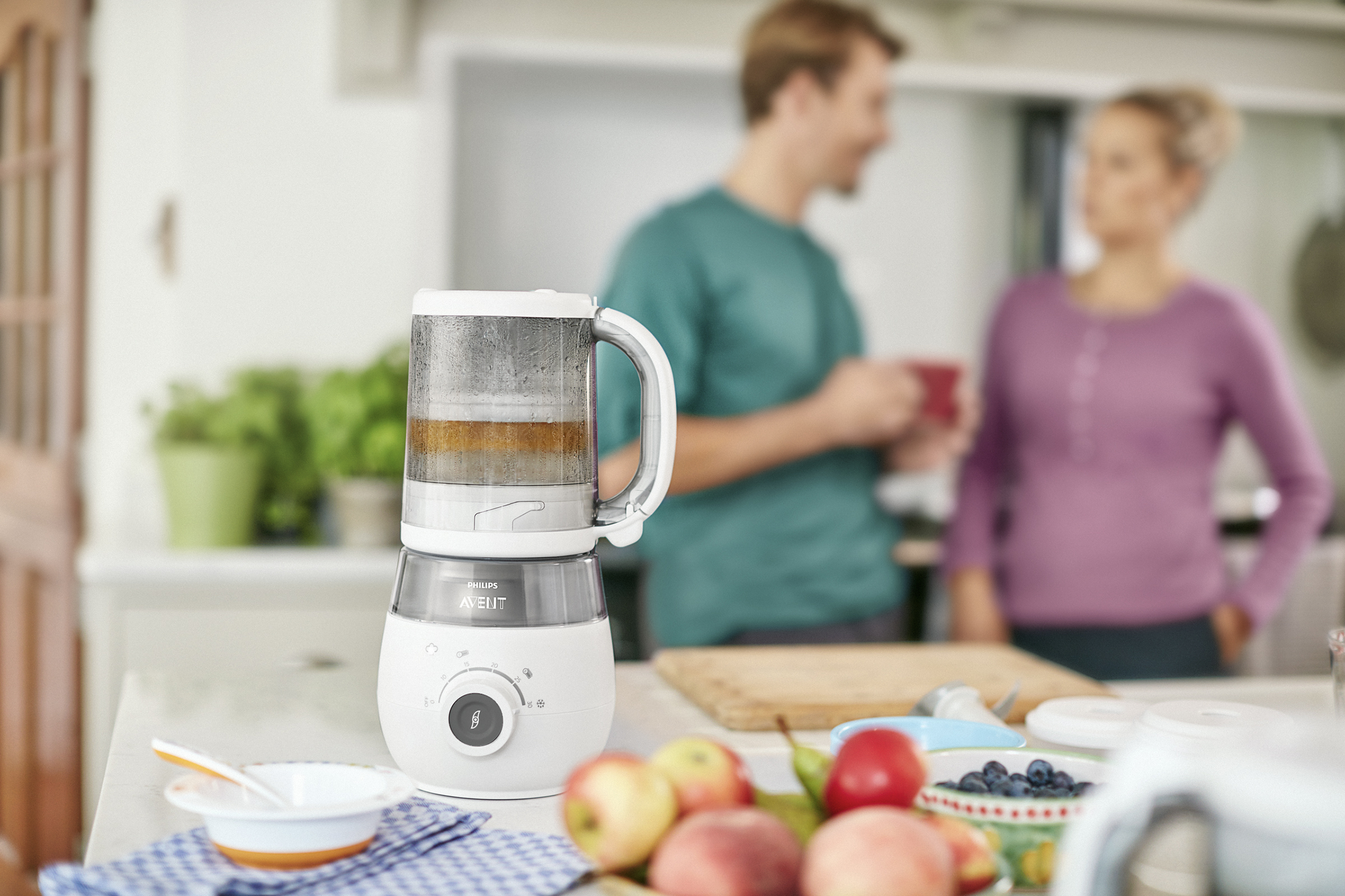 Babyvoedselmaker op een aanrecht met fruit en een stel op de achtergrond. Wit en grijs domineren de scène.