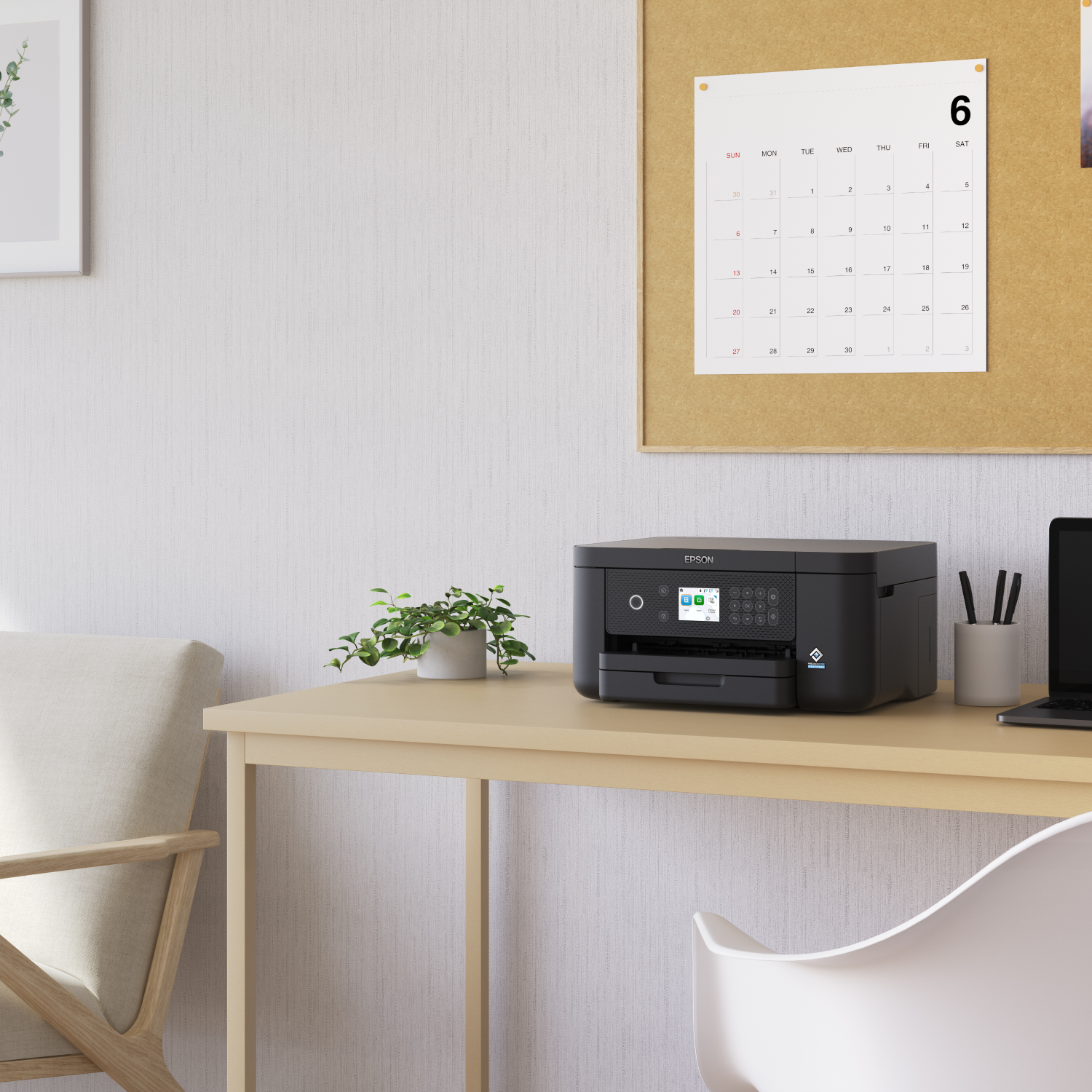 Black printer on a desk with a calendar, plant, and a laptop in the background.