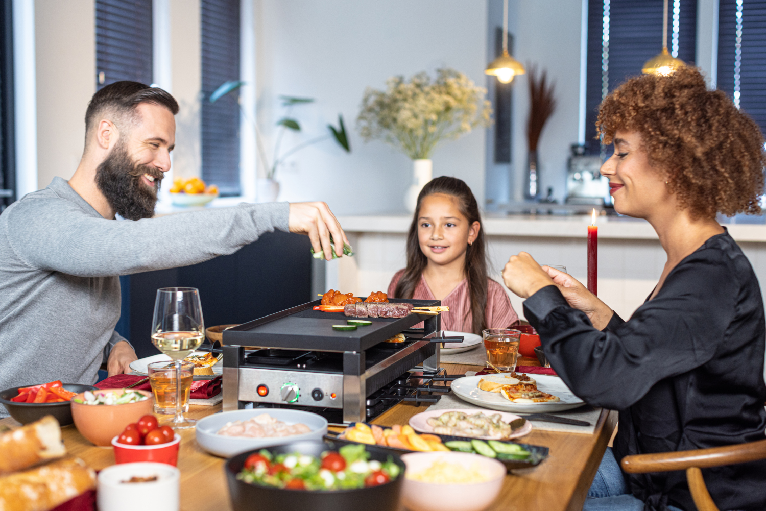 Familie rond een tafel, koken op een raclette grill. Vrolijke gezichten.