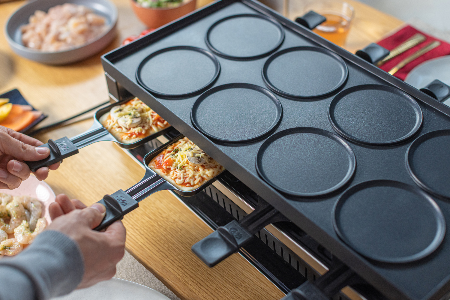 Raclette grill met kleine pizza's op peddels. Iemands handen houden de peddels vast.