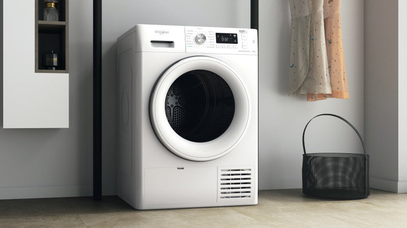 Fehér Whirlpool szárítógép egy szobában, fekete kosárral és ruhákkal. Bézs padló és falak.