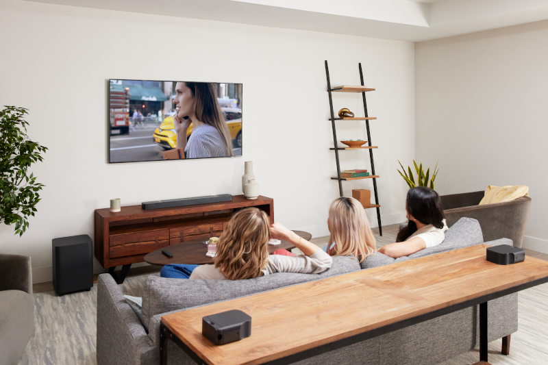 Drei Frauen schauen im Wohnzimmer fern mit Soundbar, Subwoofer und Beistelltisch.