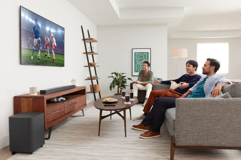 Drei Männer auf einem grauen Sofa schauen fern, eine Soundbar, ein Subwoofer und andere Heimkinoelemente sind sichtbar.