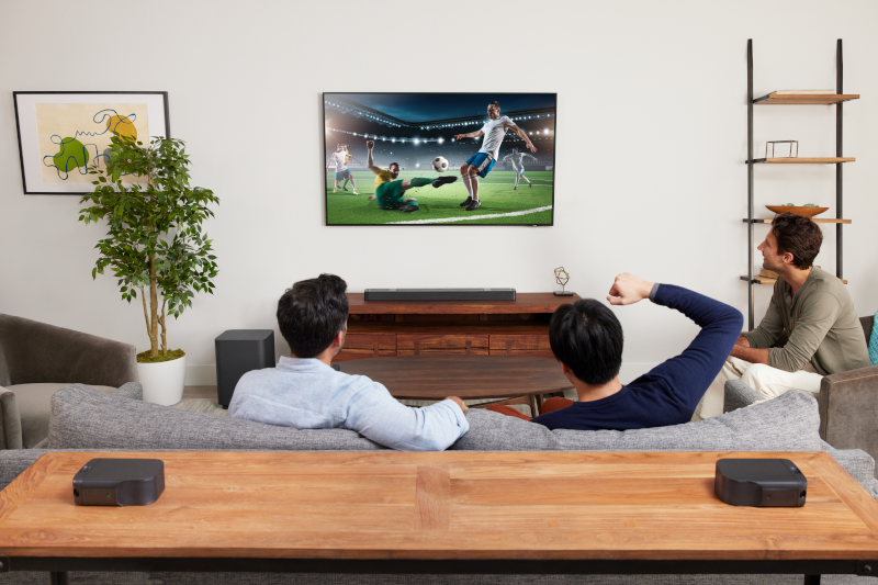 Drei Personen schauen ein Fußballspiel im Fernsehen. Der Raum hat ein Sofa, Lautsprecher und einen Holztisch.