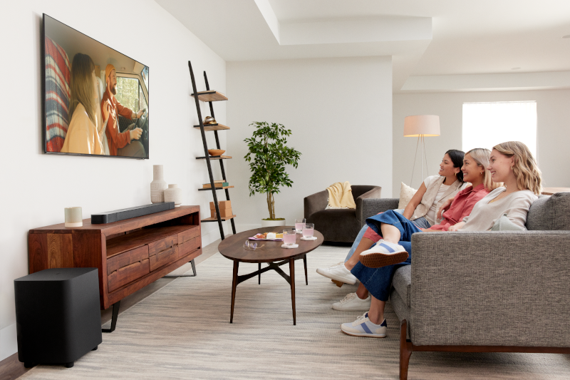 Drei Frauen auf einem Sofa sehen fern. Eine Soundbar und ein Subwoofer befinden sich vor dem Fernseher.