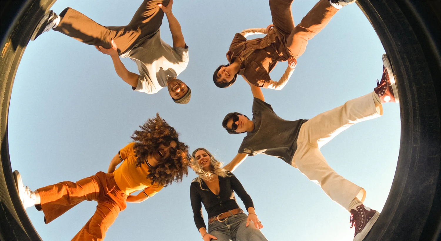 A low-angle shot of five people in a circle, looking down at the camera, with a blue sky background.