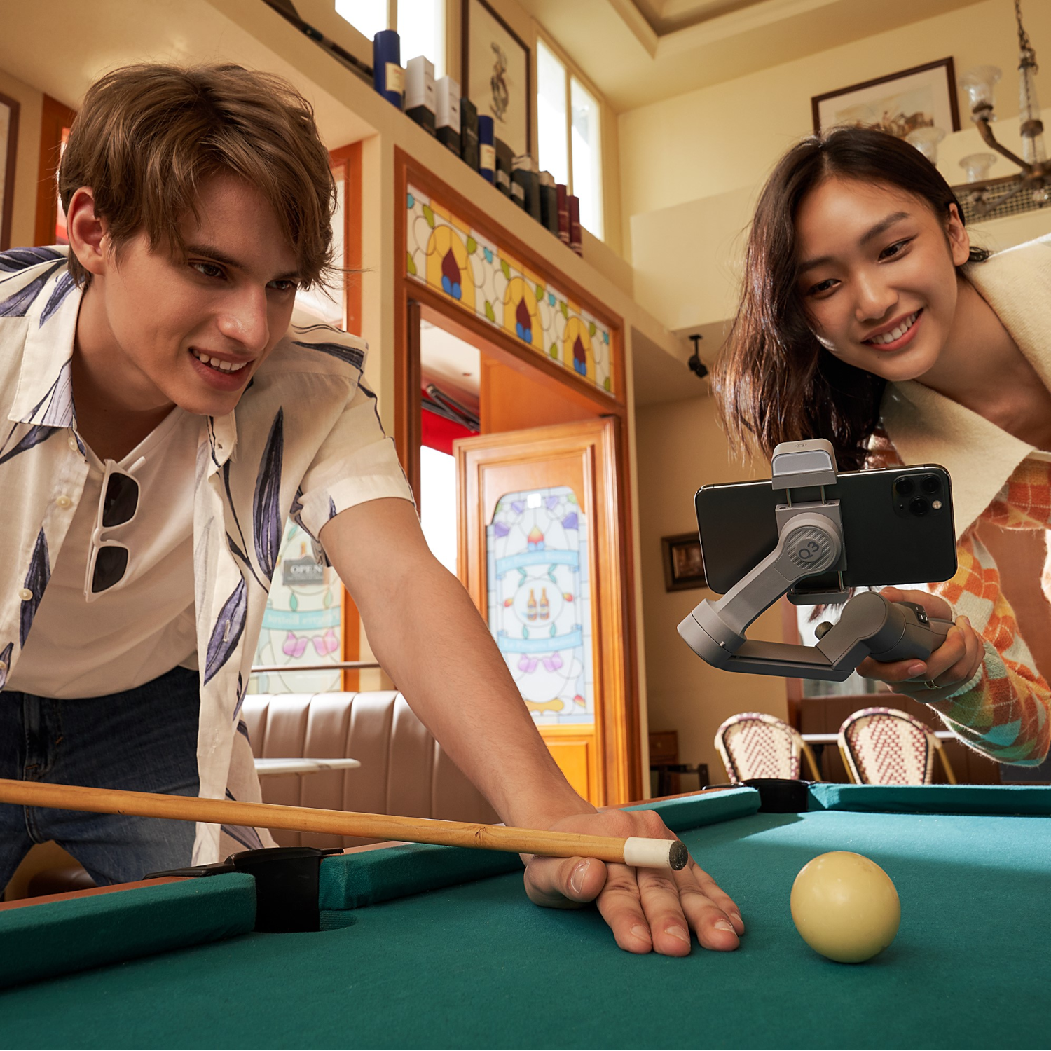 Two people playing pool with a phone on a gimbal recording them.
