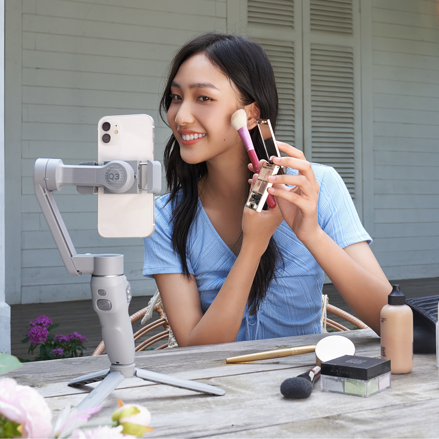 A woman applying makeup while filming with a phone on a tripod. Various makeup items are on the table.