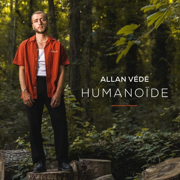 Homme debout sur des souches de bois dans une forêt, portant une chemise orange. Texte : Allan Vedé Humanoid.