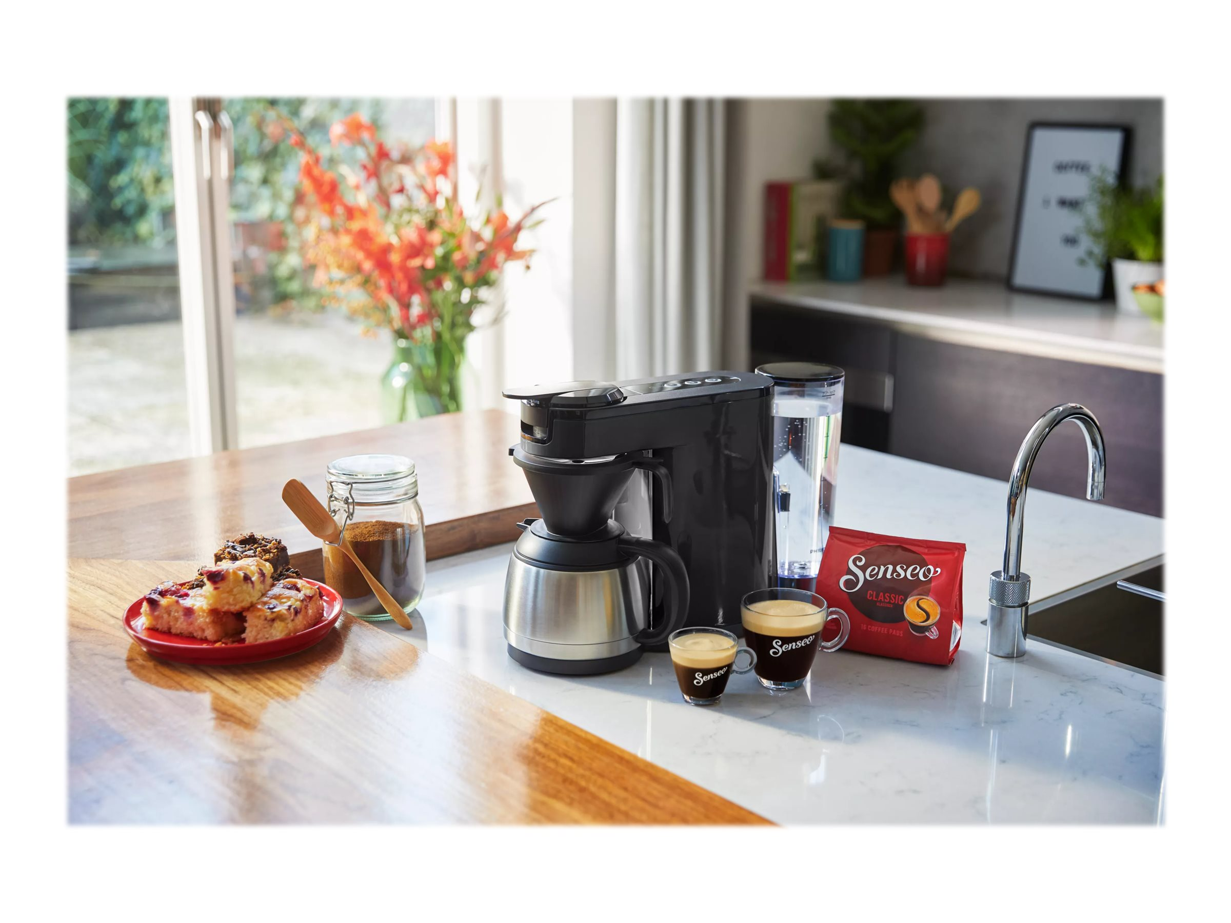 Kaffeemaschine mit Zubehör auf einer Theke. Kaffeetassen, Kaffeebohnen und Gebäck sind vorhanden.