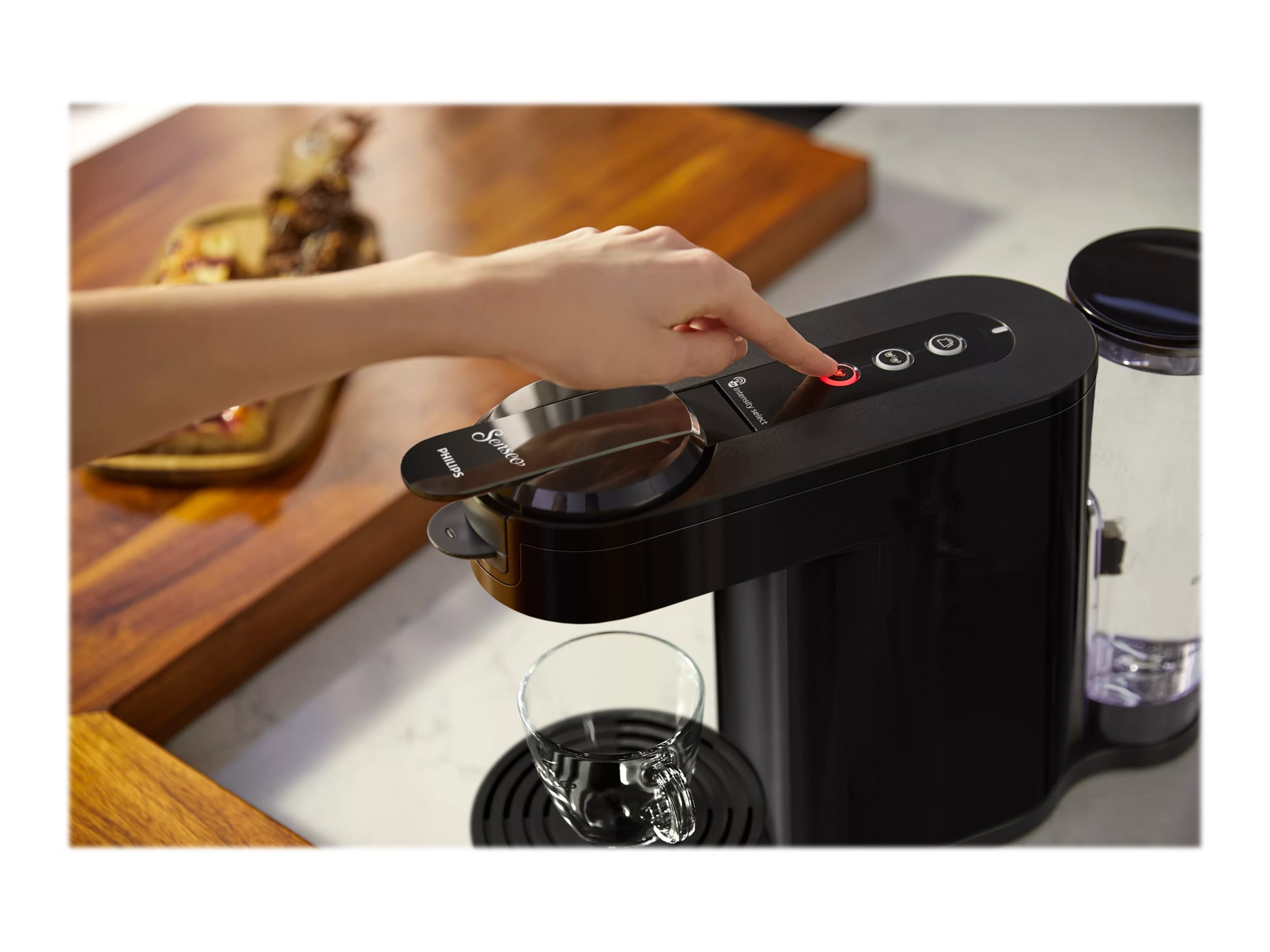 Schwarze Kaffeemaschine. Eine Hand drückt den Einschaltknopf. Ein Glas darunter.
