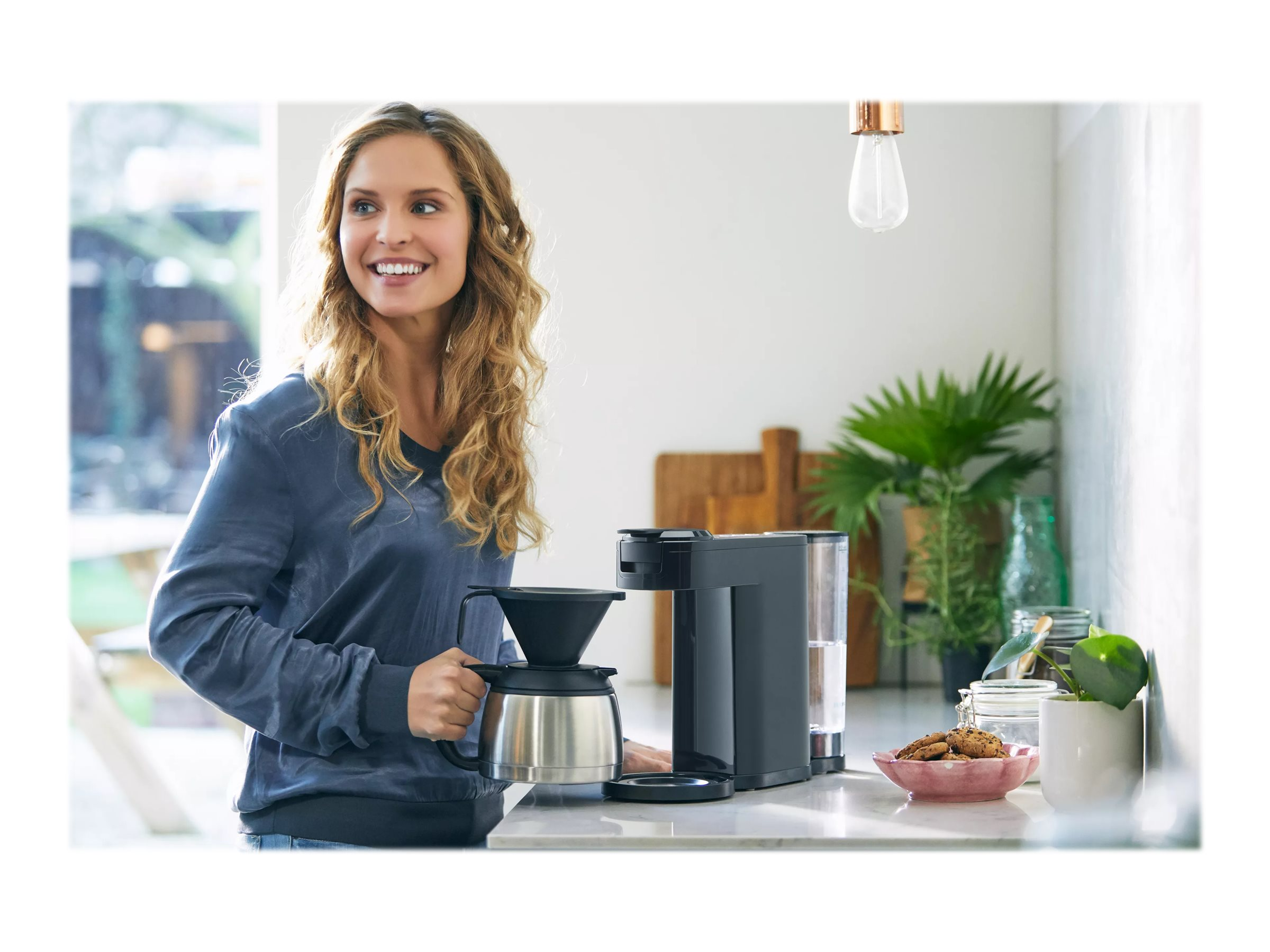 Frau hält Kaffeemaschine, lächelt, in Küche mit Pflanze und Keksen auf Teller.