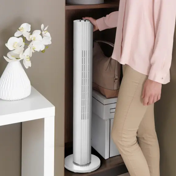 Hoge witte ventilator in een kast. Een vrouw reikt ernaar. Een witte vaas en bloemen staan op een tafel.