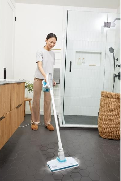Une femme utilisant un balai vapeur blanc dans une salle de bain, avec une douche et des meubles en bois visibles.