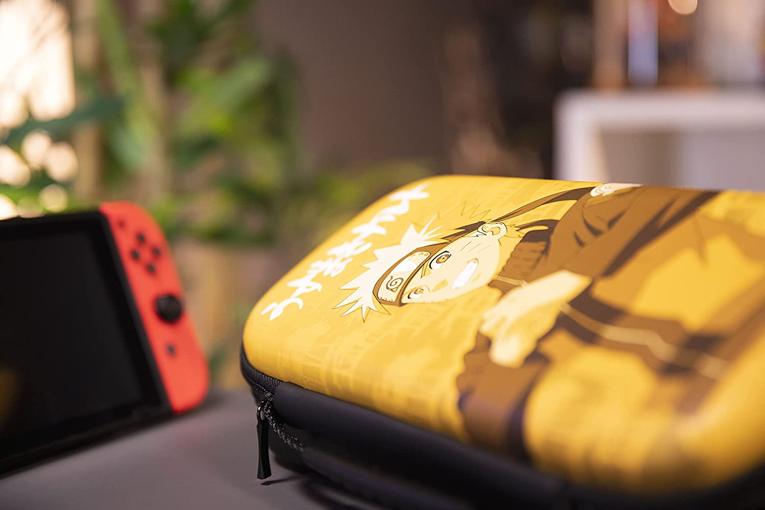 Custodia Switch Naruto gialla accanto a una console Switch rossa. Sfondo sfocato, con una pianta.
