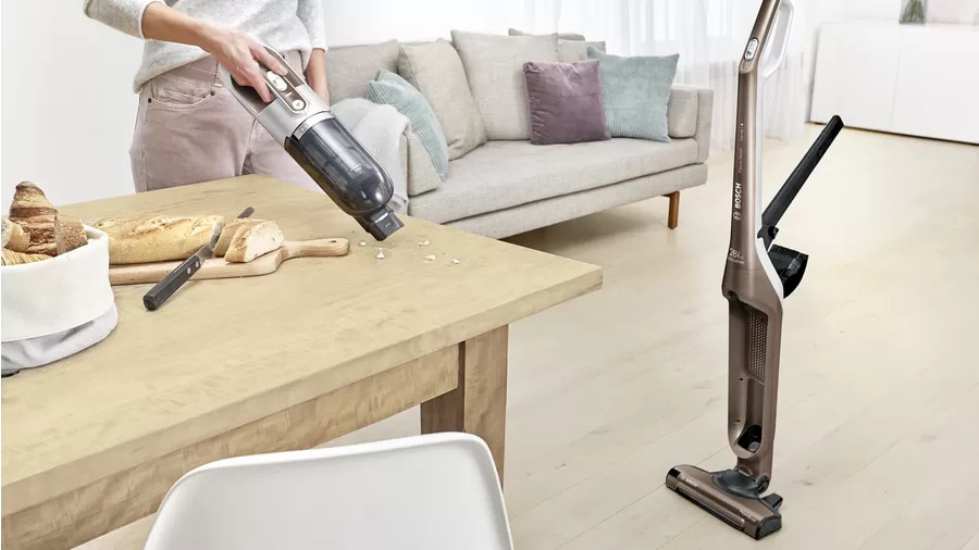 Een persoon stofzuigt kruimels van een tafel. Een staande stofzuiger staat rechts.