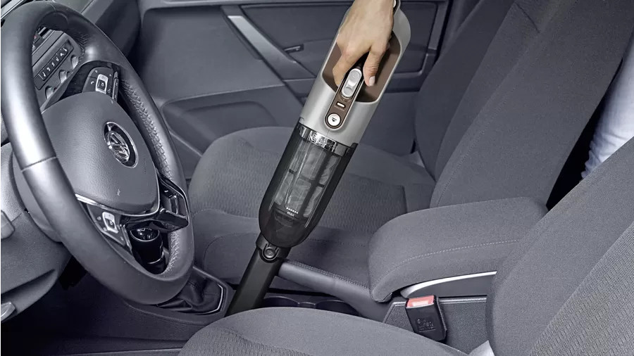 Een hand stofzuigt een auto-interieur. De stofzuiger is grijs en zwart. De autostoelen zijn grijze stof.