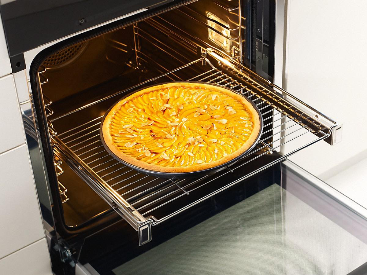 Ofen mit Tarte auf einem Rost. Die Tarte ist rund mit gelbem Obst und Nüssen. Ofentür ist offen.