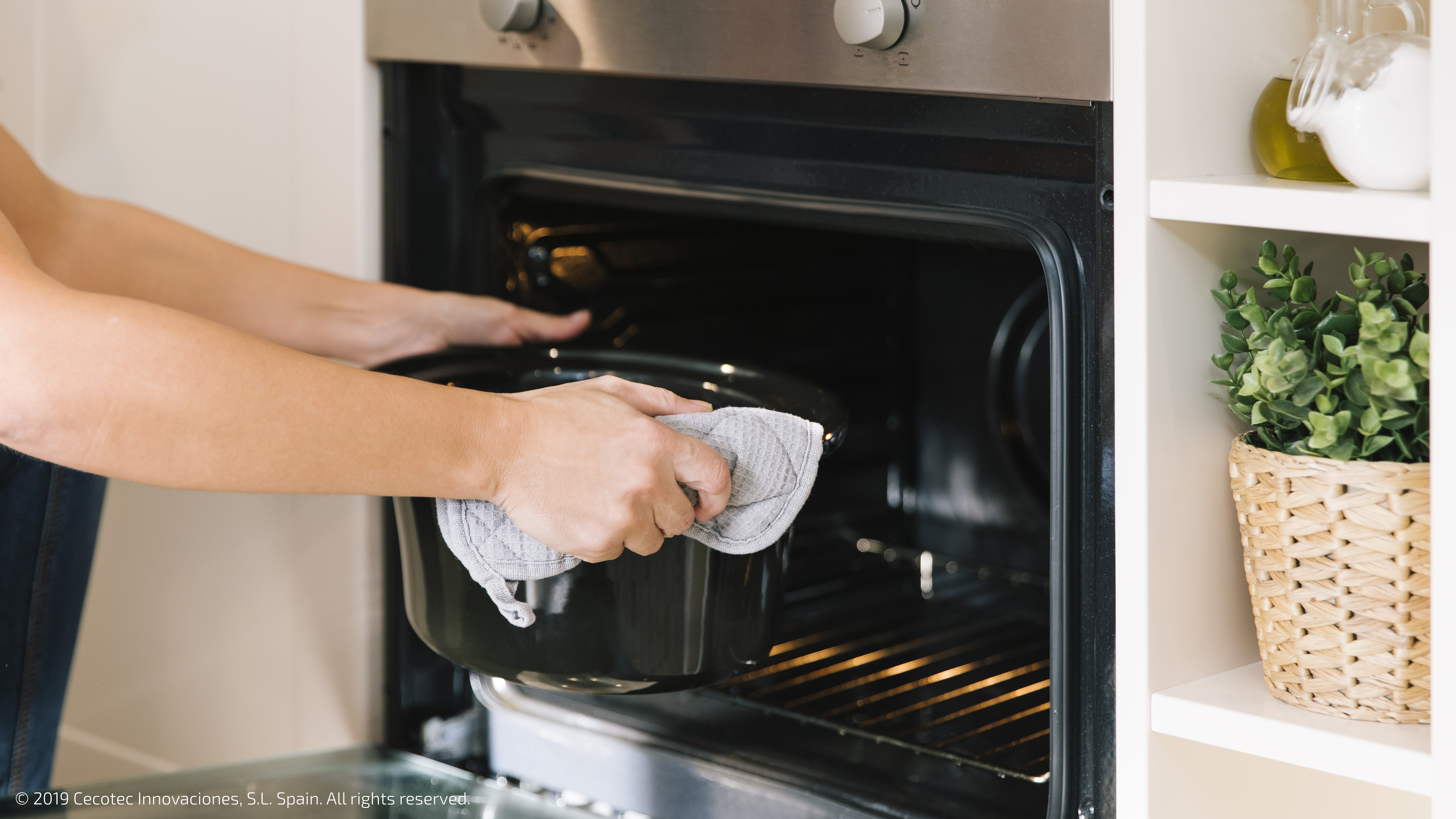 Manos colocando una fuente para hornear negra en un horno caliente, con una estantería de cocina en el fondo.
