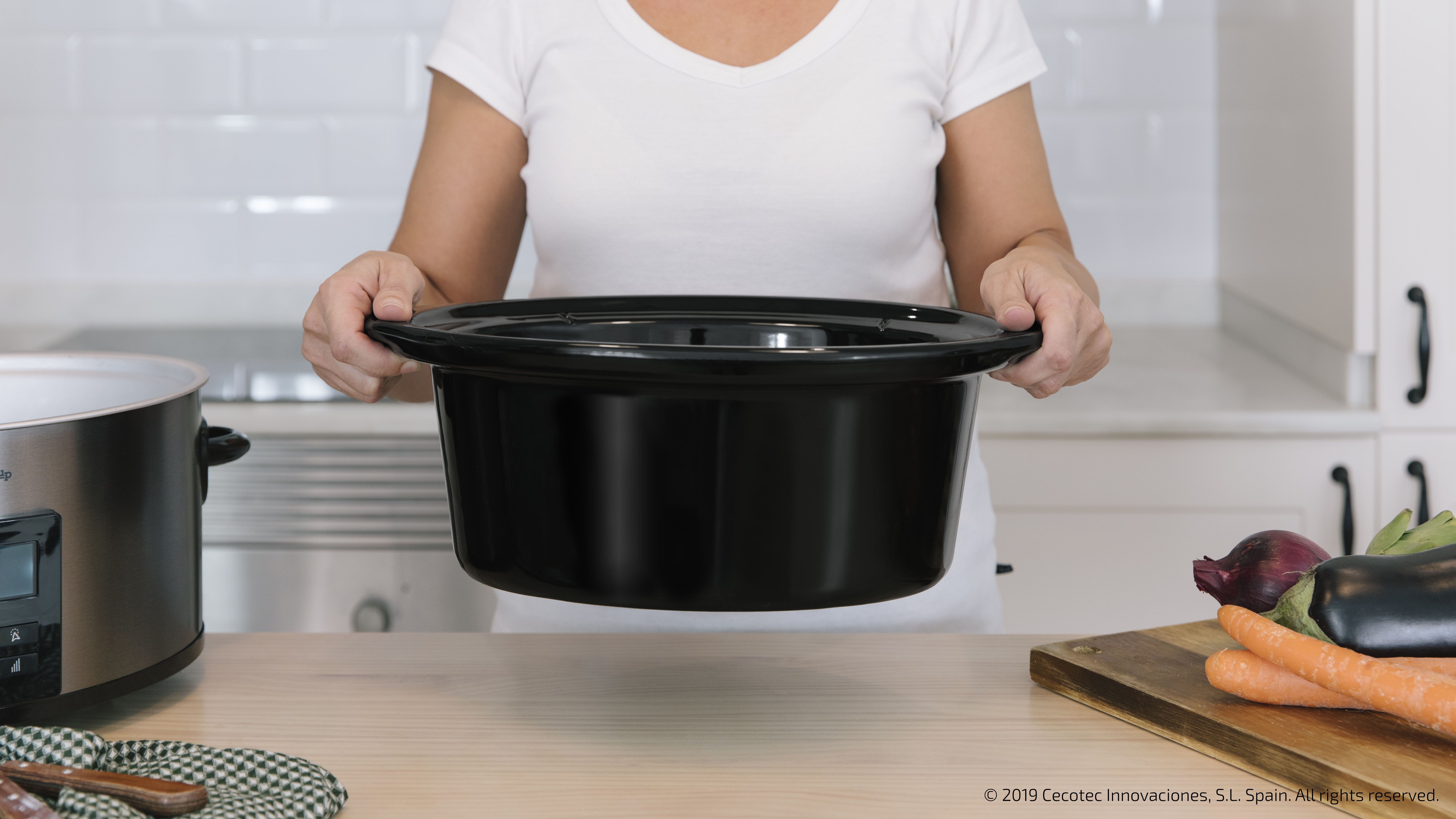 Una persona sostiene una olla de cocción lenta negra. Verduras y una tabla de cortar en la mesa.