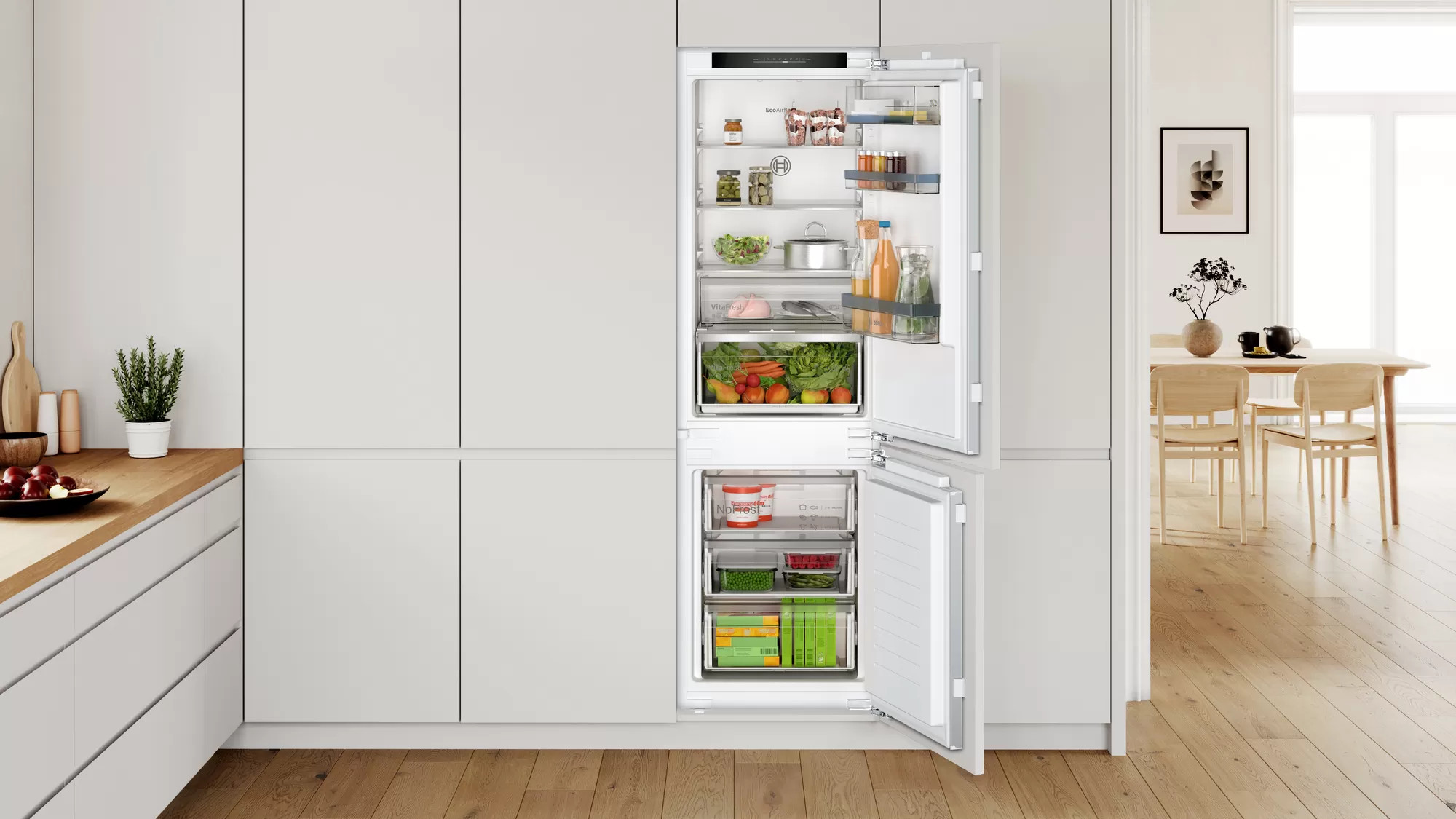 Un réfrigérateur ouvert avec des aliments et des légumes visibles à l'intérieur, dans une cuisine blanche.