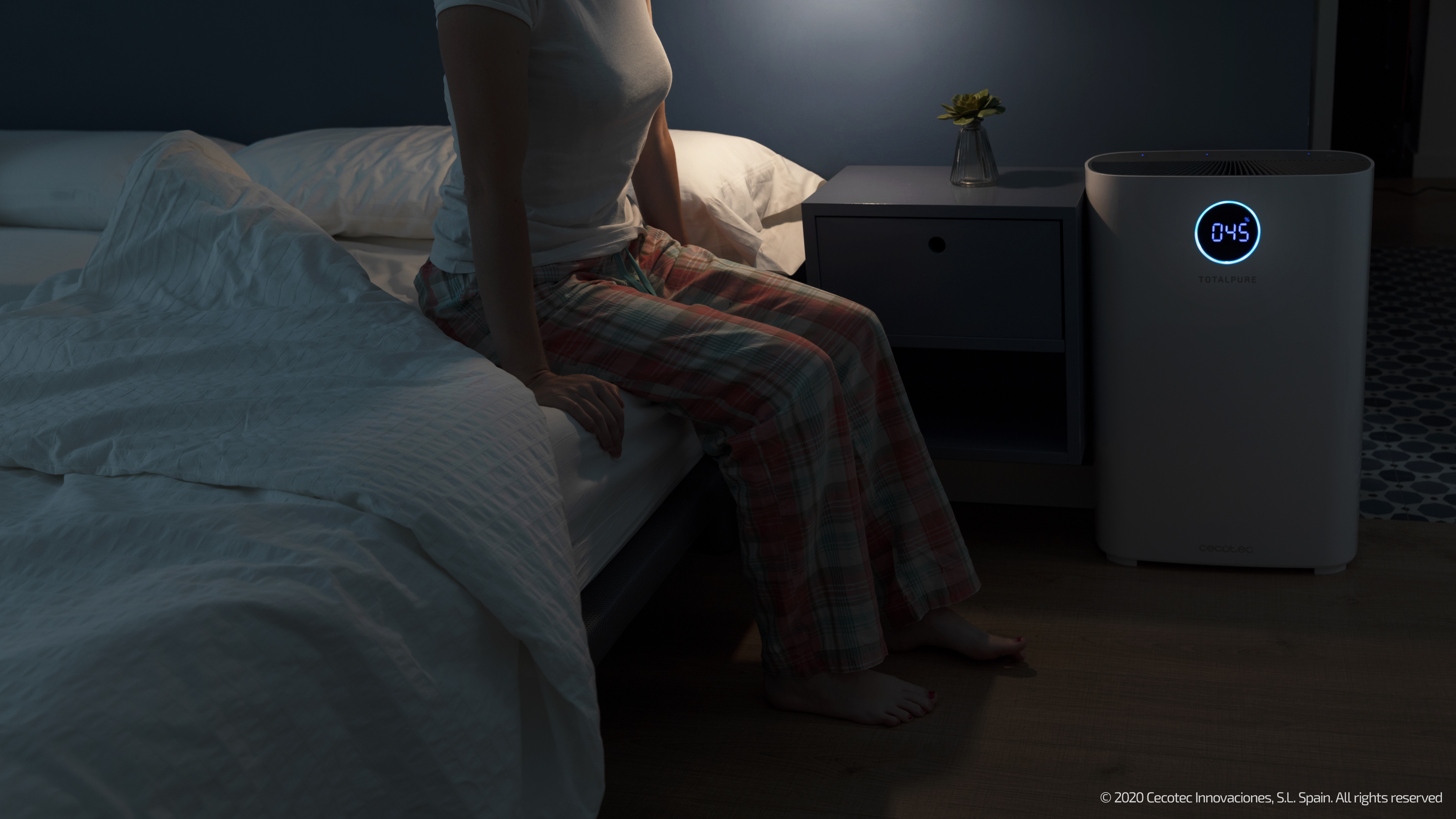 Una mujer sentada en una cama, un purificador de aire a su lado. La escena está poco iluminada.