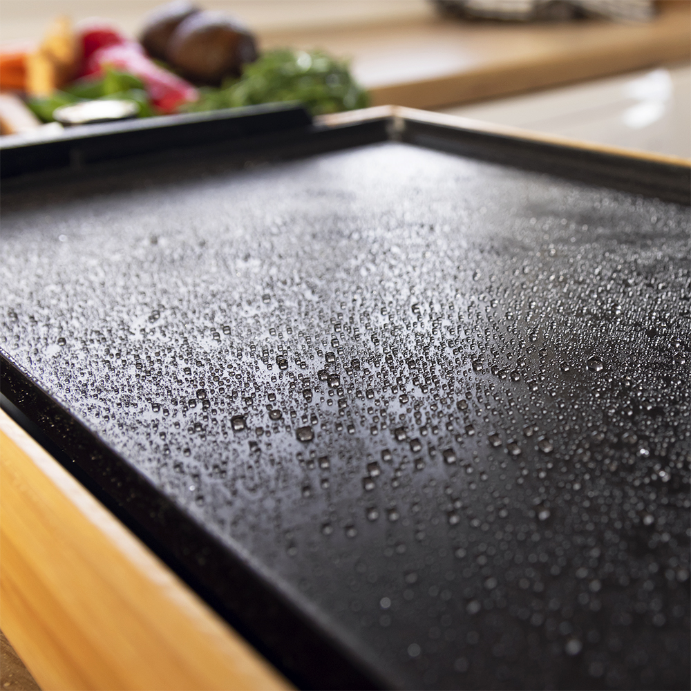 Plancha negra con gotas de agua. Marco de madera y verduras borrosas en el fondo.