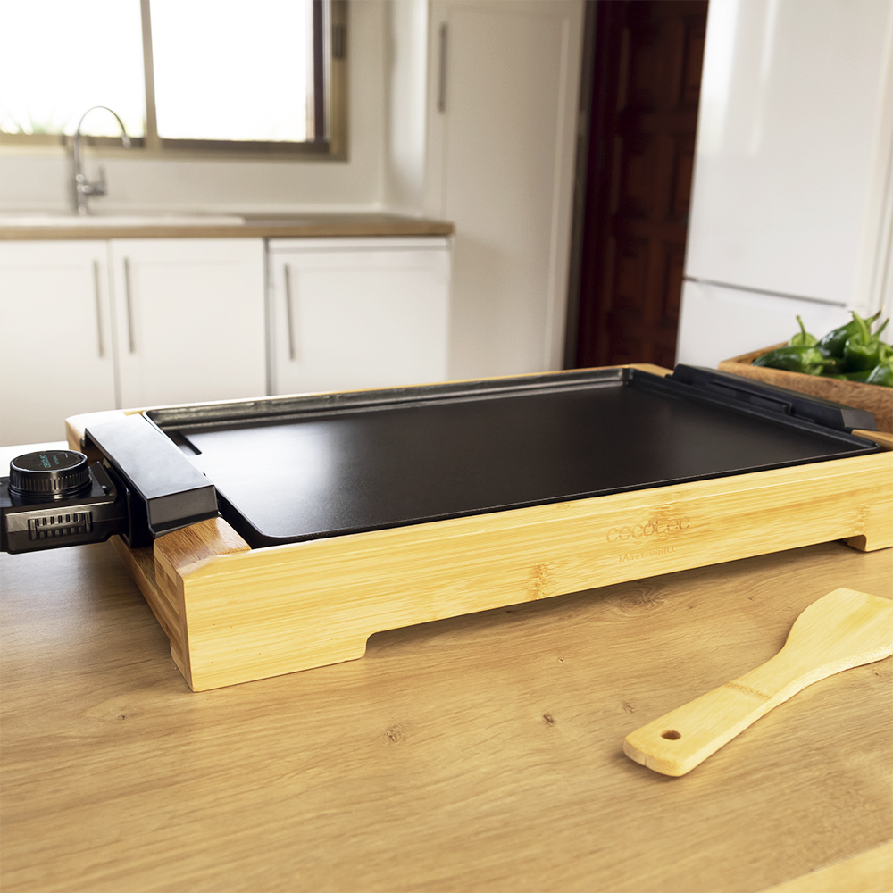 Una plancha negra sobre una base de madera. Una espátula y verduras en la encimera.