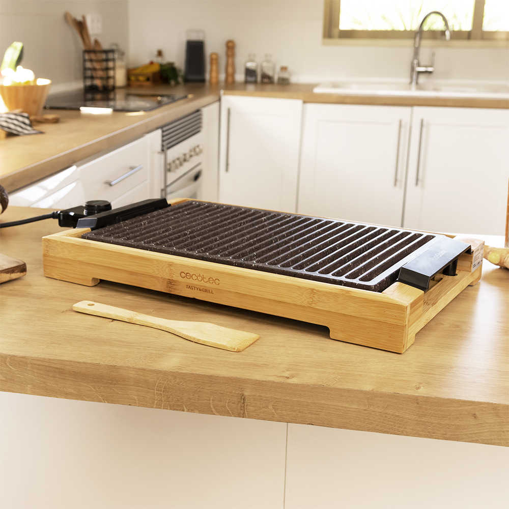 Una parrilla eléctrica de madera sobre una encimera con una espátula, en una cocina luminosa.