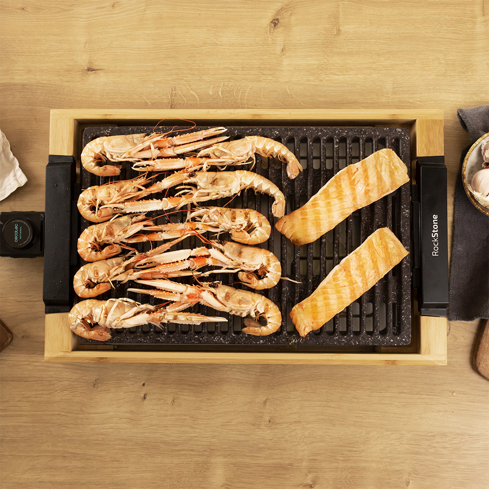 Mariscos y salmón a la parrilla en una parrilla negra con marco de madera, sobre una mesa de madera.