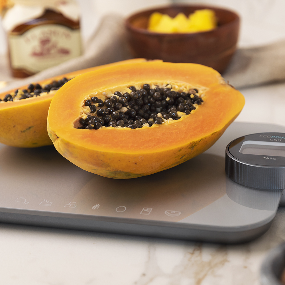 Mitades de papaya en una balanza. Semillas negras visibles. Cuenco y tarro de madera en el fondo.