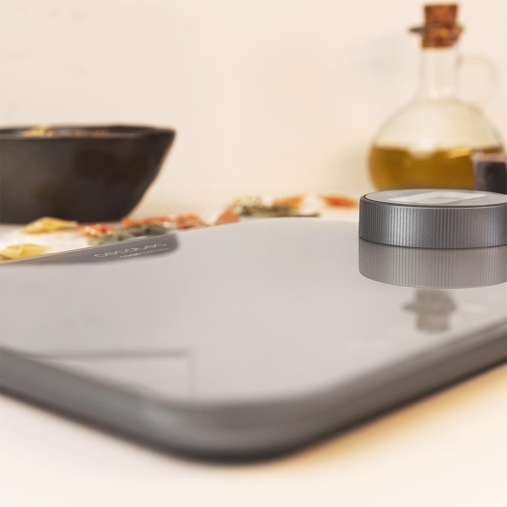 Una báscula de cocina e ingredientes. Báscula gris, cuenco negro, botella de aceite y cuchillo. Fondo beige.