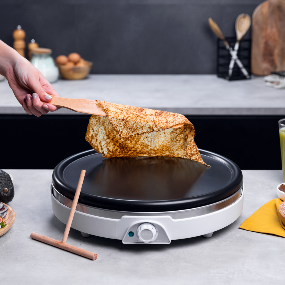 Una mano voltea un crepé en una plancha con utensilios de cocina cerca.