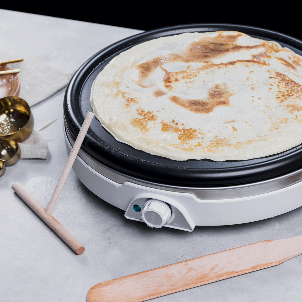 Un crepe que se cocina en una plancha eléctrica blanca, con accesorios sobre la mesa.