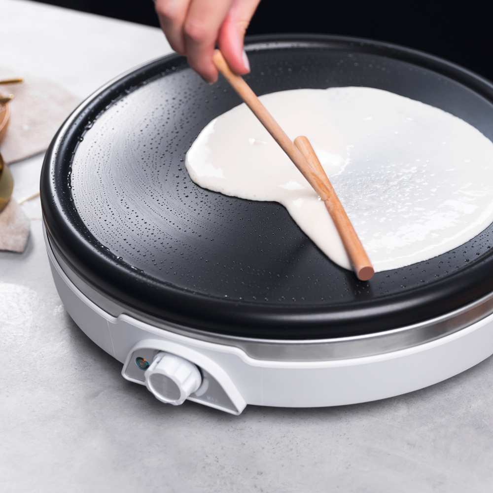Una persona usa una herramienta de madera para extender la masa en una plancha caliente. Primer plano.