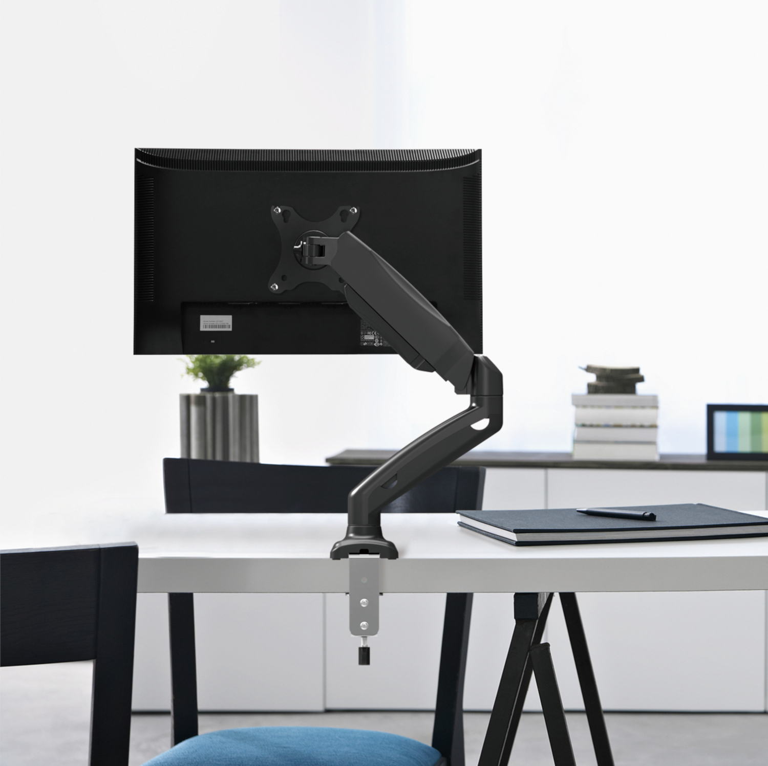 Monitorarm met een zwarte monitor op een witte bureau, tegen een witte muur.