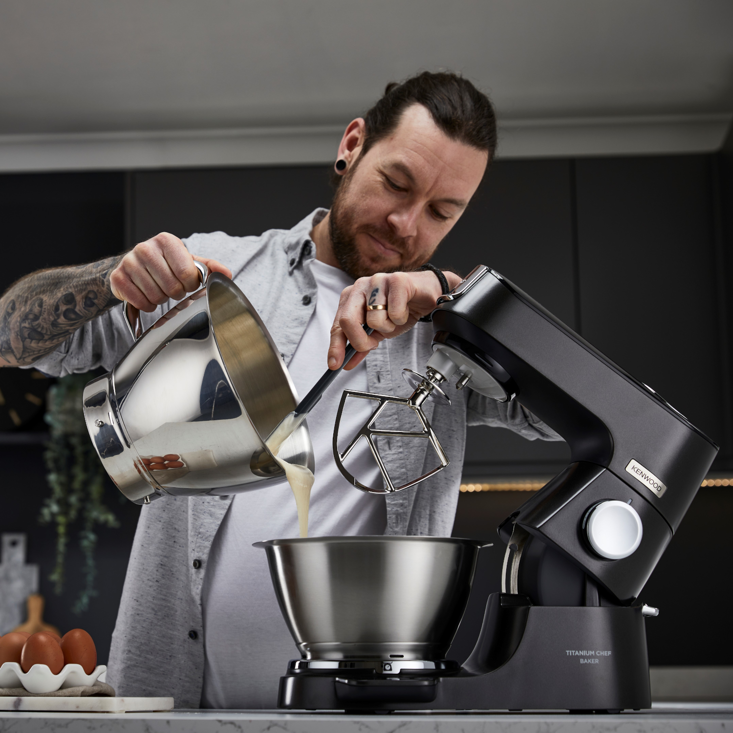 A man pouring batter into a bowl with a mixer. There are eggs on a counter.