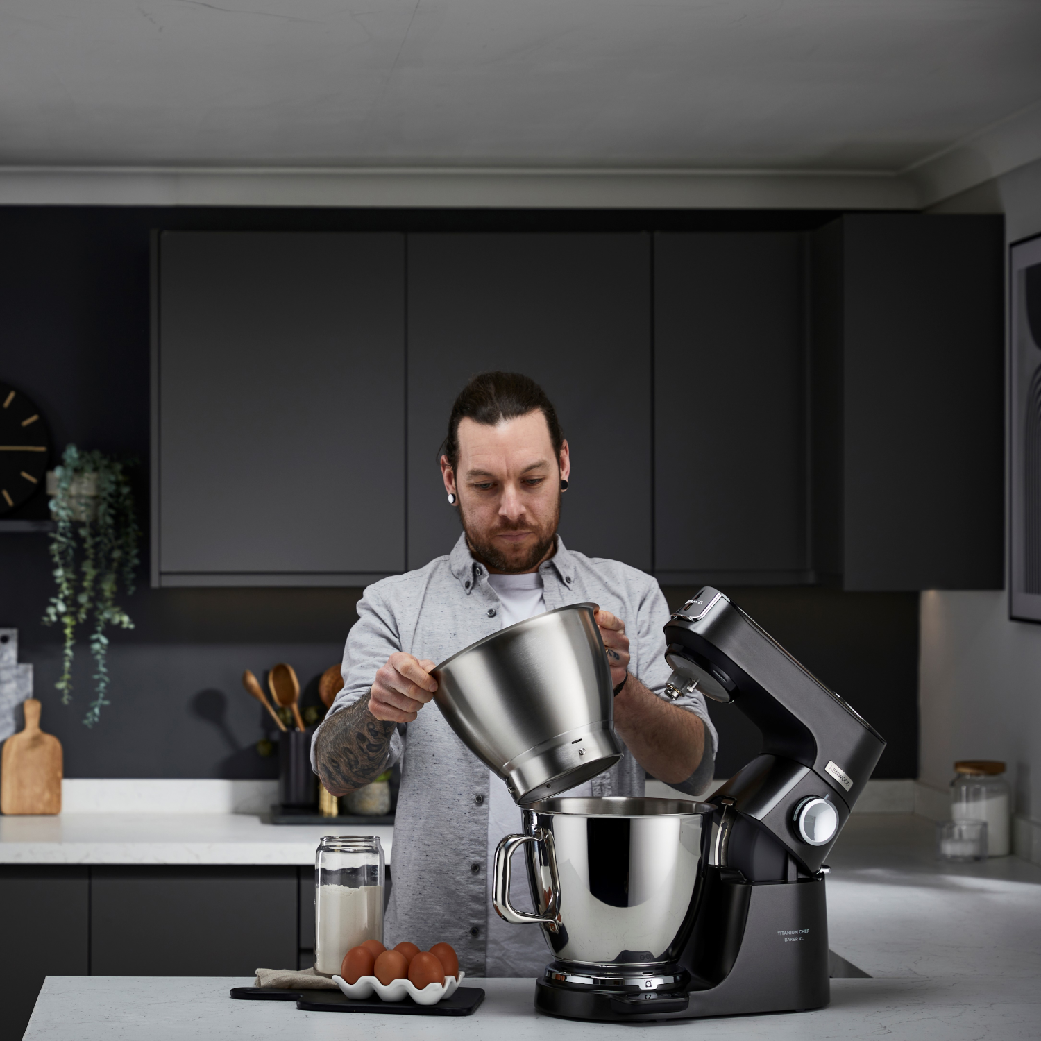 A man pours ingredients into a black stand mixer. Kitchen setting.