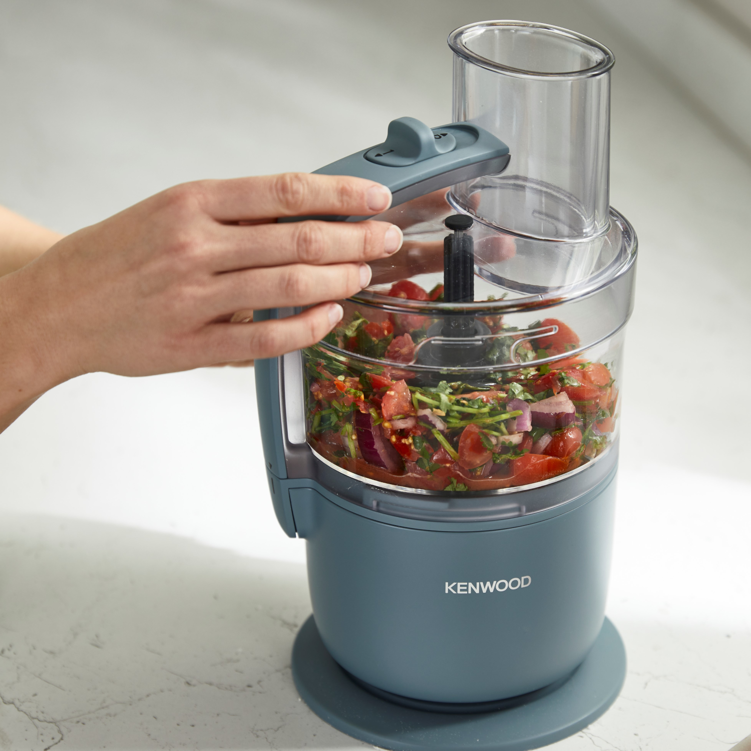 A person's hands using a food processor. The processor is teal, with chopped tomatoes, herbs and onions inside.