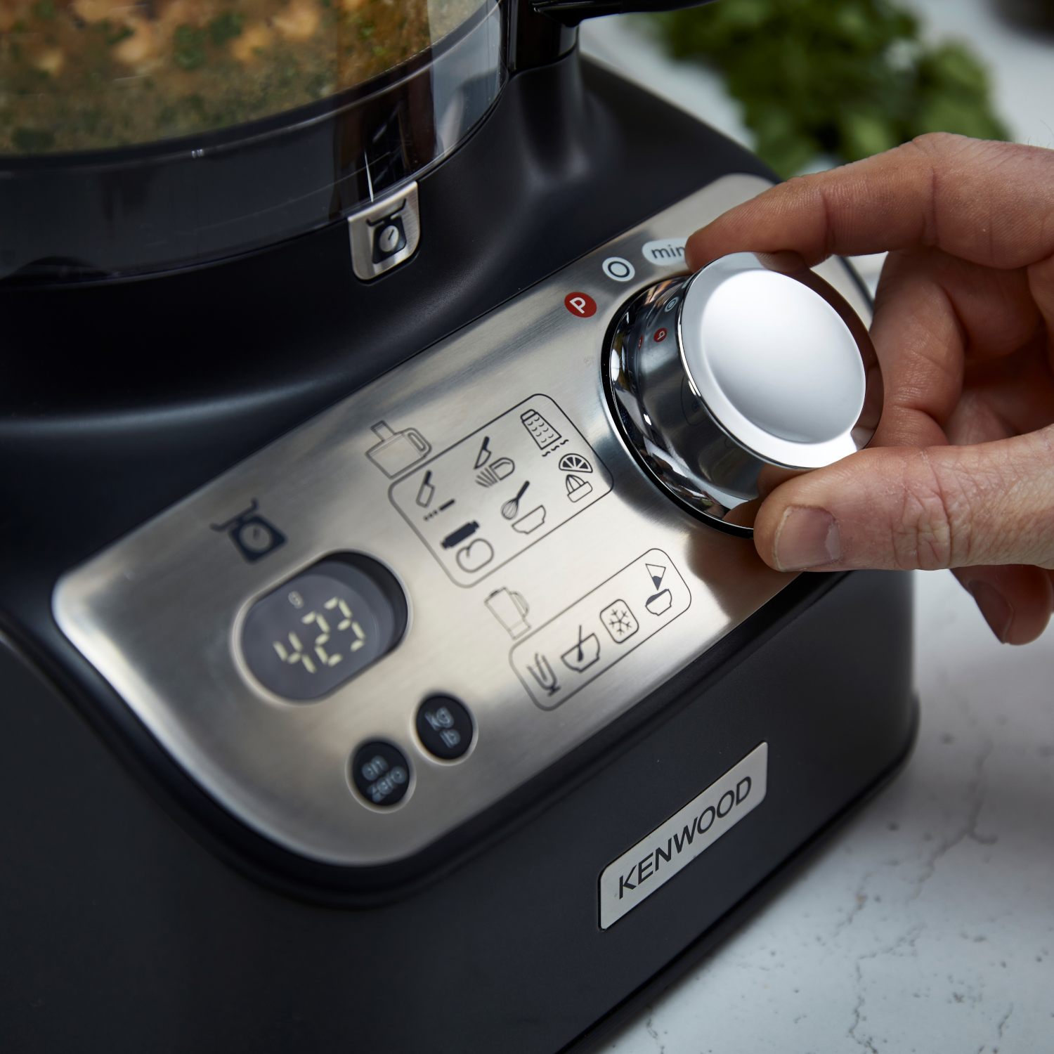 A hand turns a dial on a Kenwood food processor. The dial has settings and a digital display reads '4:23'.