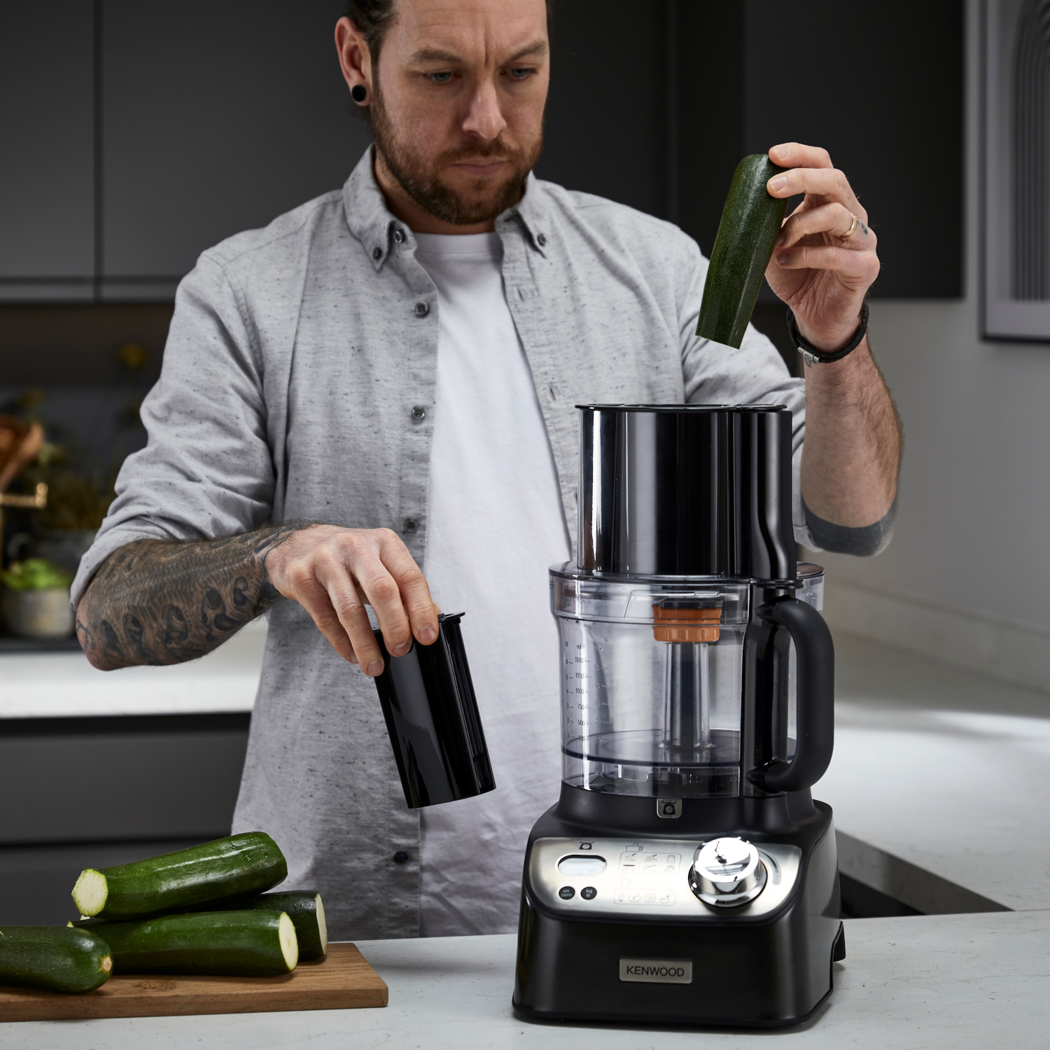 A man prepares food with a food processor. Zucchinis are on the counter.
