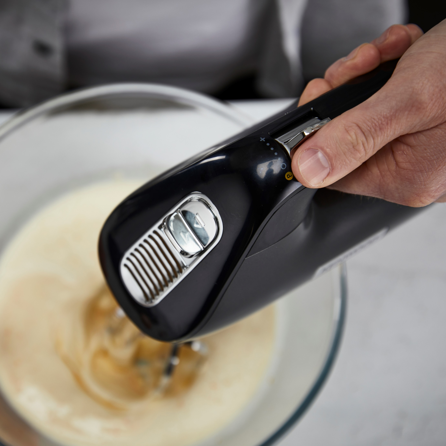 A person's hand holding a black hand mixer over a glass bowl with creamy mixture. The mixer is running.