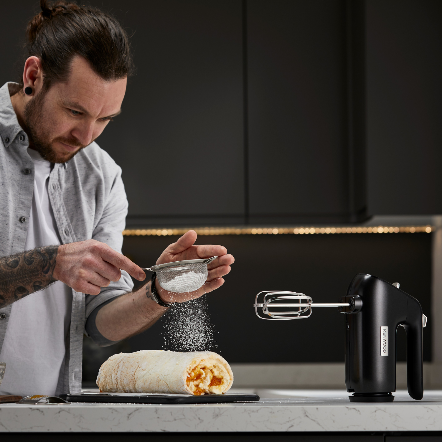 A man sprinkles powdered sugar on a roll cake with a hand mixer in a modern kitchen setting.