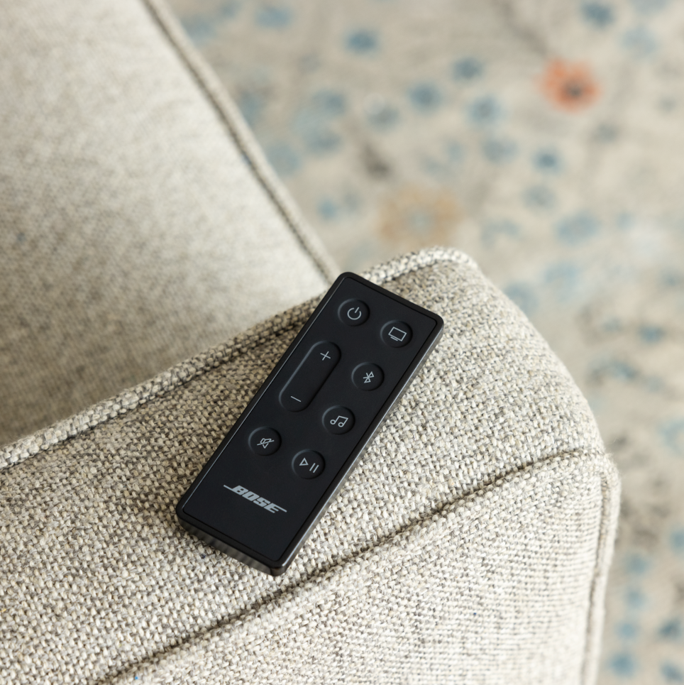 Zwarte Bose-afstandsbediening op een grijze, getextureerde fauteuil. Knoppen voor stroom, volume, muziek, etc.